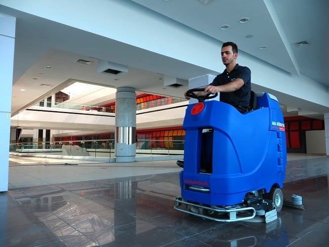 An operator driving a blue floor scrubber through a large indoor open space